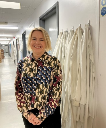 Joanna at work in the labs, standing in front of a row of lab coats on coat hooks.