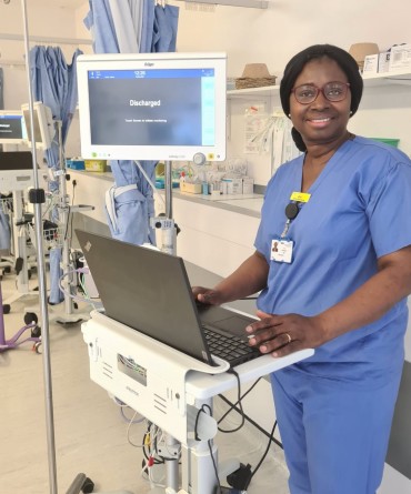 Abimbola Ojo in blue scrubs. She is stood next to a computer on a stand and in front of medical equipment on a ward.