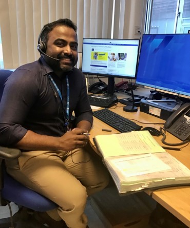 Ribu sitting at his desk which has two screens and a large open book. He is wearing a phone headset, looking ready to pick up a call!