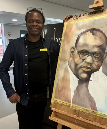 Patient Services Assistant Nduka Omeifa standing next to a portrait that he has painted