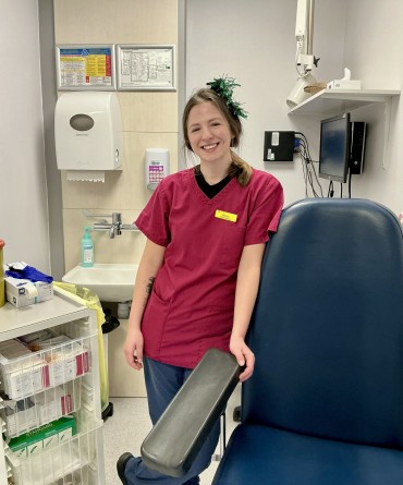 Rebecca is in red scrubs, standing beside a blue examination chair in a medical room with supplies and equipment. She has green tinsel in her hair.
