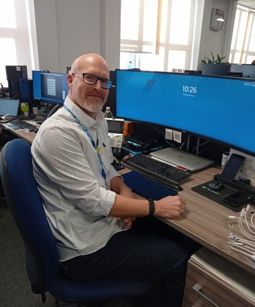 A man with glasses, wearing a white shirt, sits at a desk with multiple computer monitors in an office. He looks at the camera and smiles.