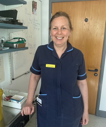 A nurse in her uniform smiling. She is standing in a consultant's office with clinical equipment in the background.