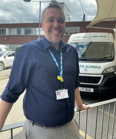Joe standing against a railing outside the front of York Hospital. There is a patient transport ambulance behind him.