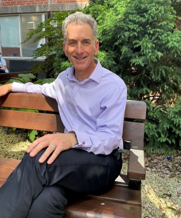 A smiling man in a blue shirt sits on a bench in a garden