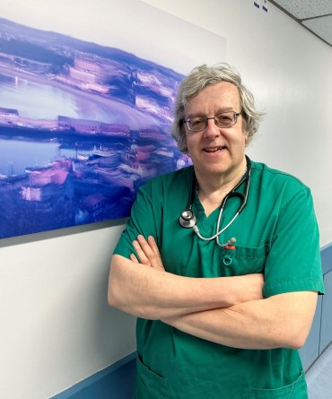 David Humphriss wearing green scrubs and a stethoscope around his neck, standing next to a painting of Scarborough beach hung on a wall in a corridor.
