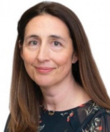 A woman with long brown hair wearing a dark patterned blouse, smiling softly at the camera against a plain white background.