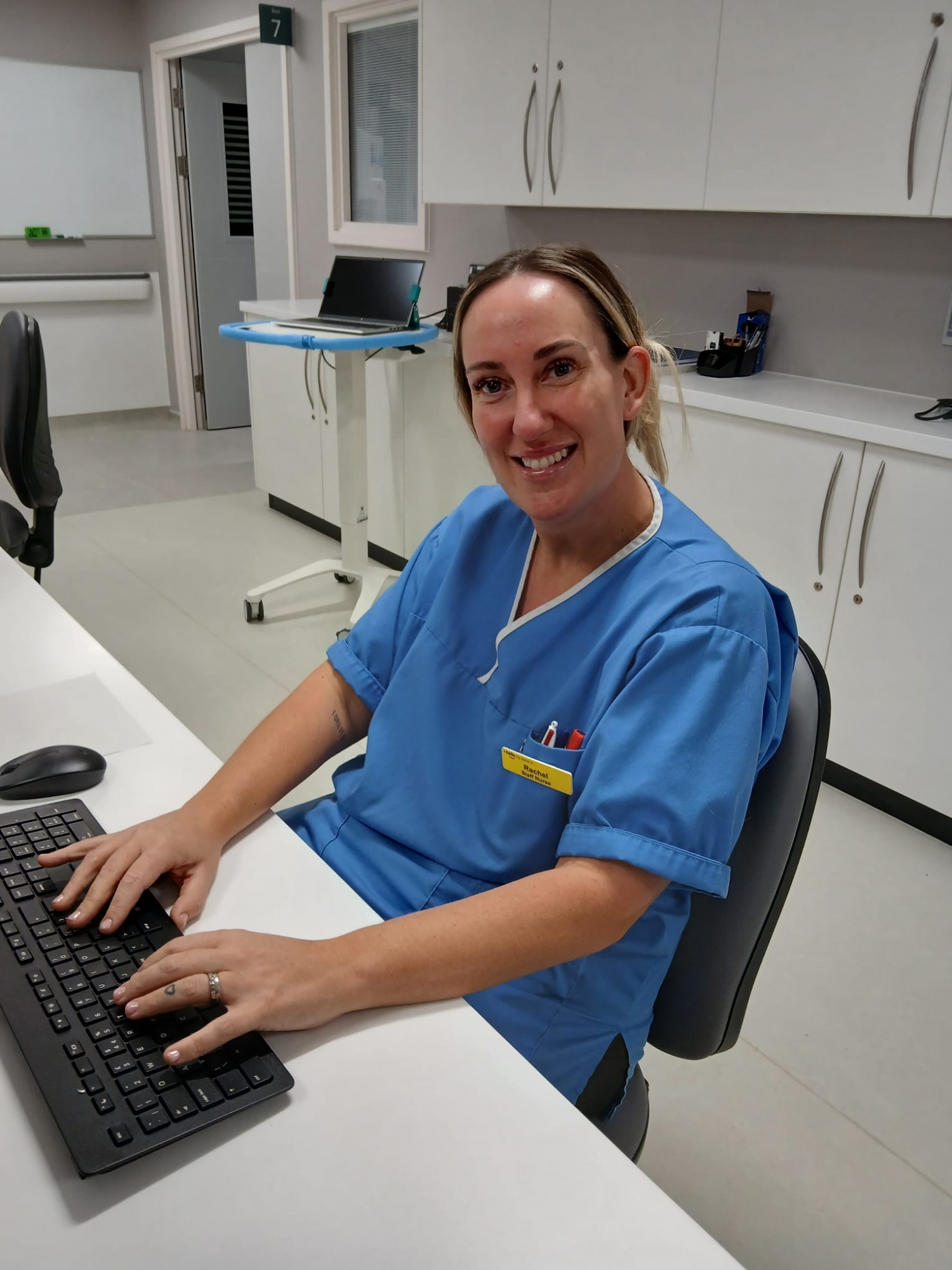 Rachel Wilson, Staff Nurse, in blue scrubs sitting at a desk typing on a keyboard.