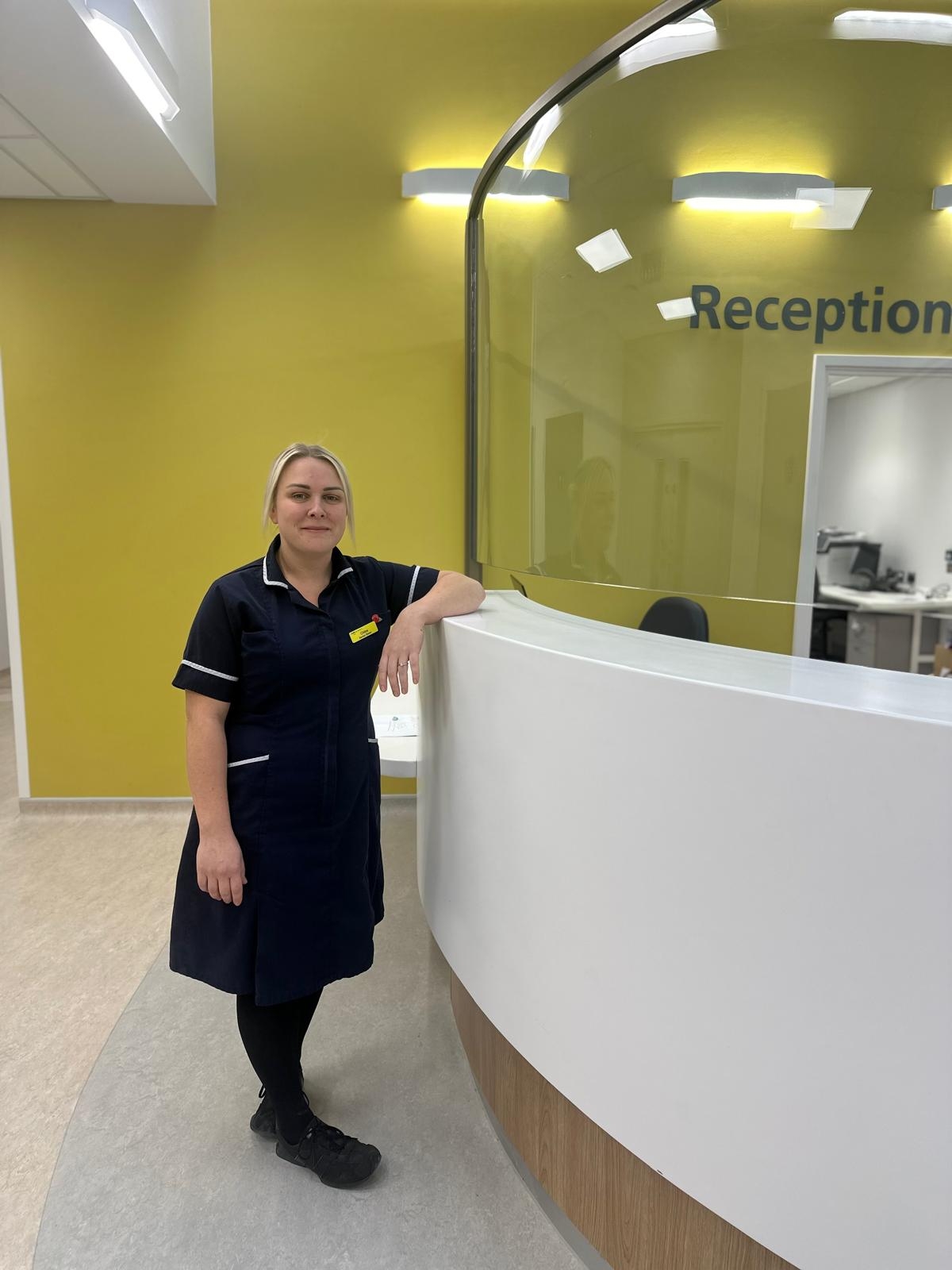 Chloe Mason, Senior Sister, wearing a navy blue nursing uniform, standing next to a reception desk.